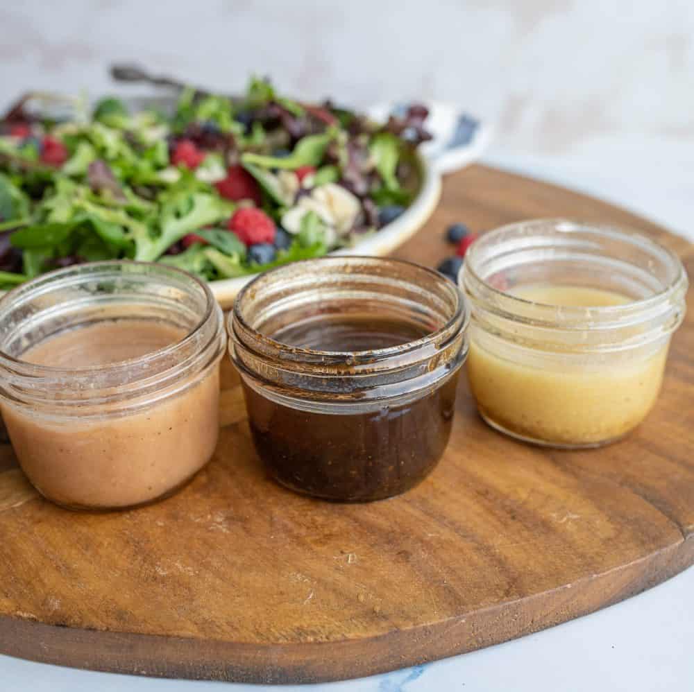 three small jars with dark and light salad dressings in them on a brown board with salad on plate in background.