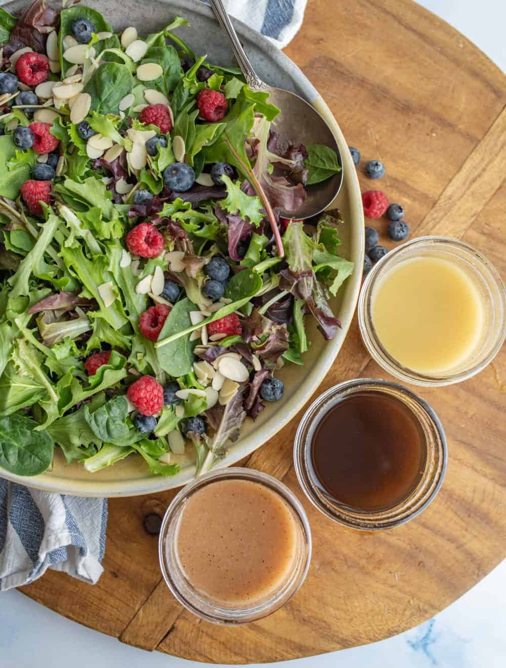 three small jars with dark and light salad dressings in them on a brown board with salad on plate.