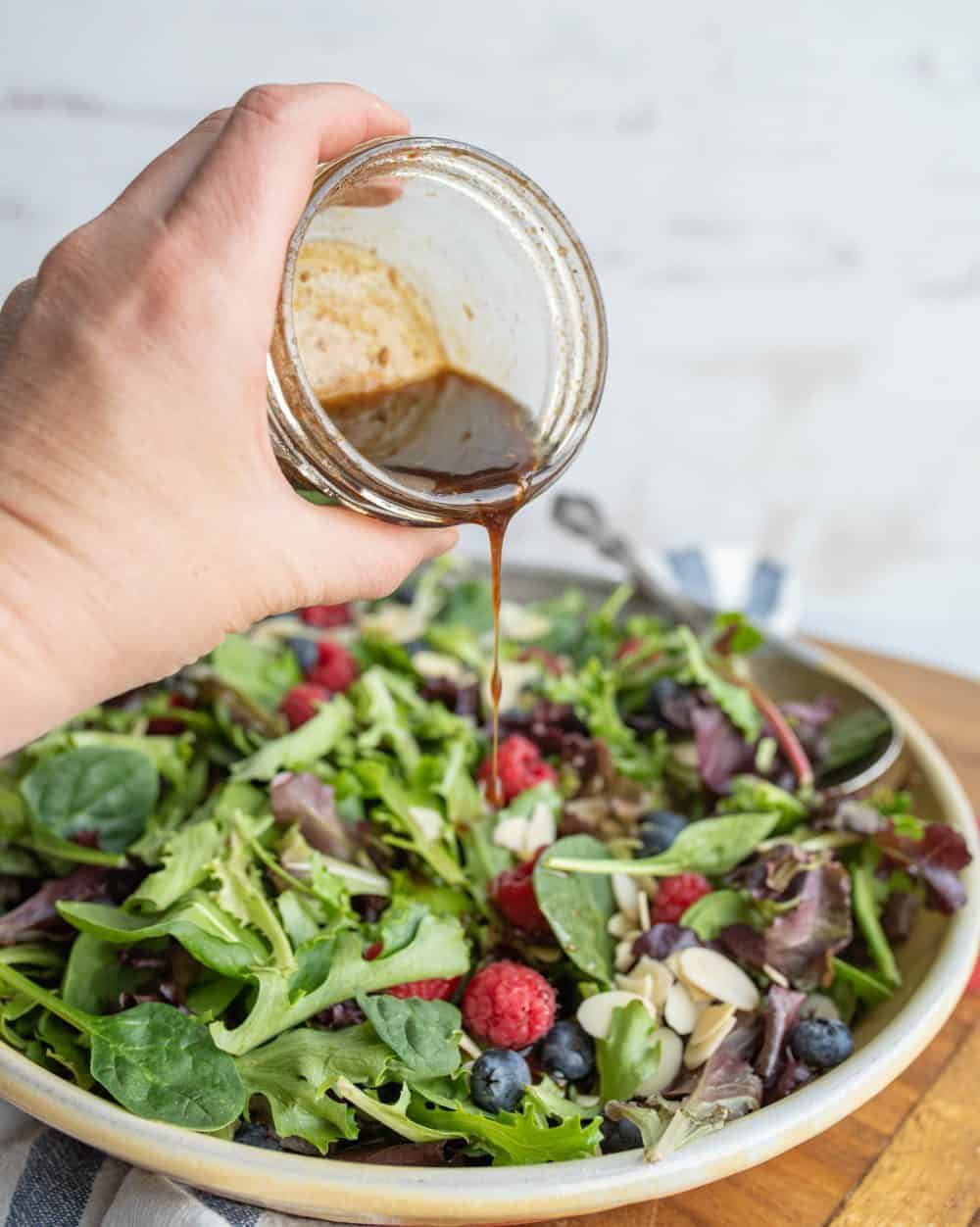 green salad on a plate with red and blue berries with a hand pouring dressing out of a jar onto salad.