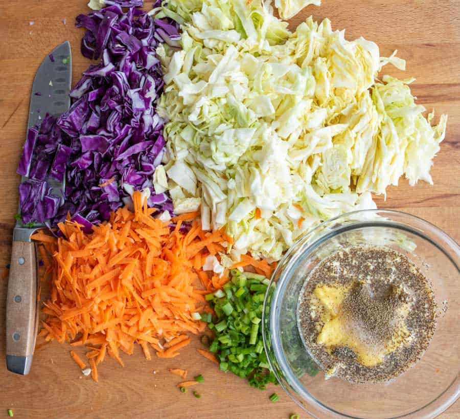 clear bowl with coleslaw dressing ingredients un-mixed and all of the raw veggies around it including carrot - white and purple cabbage - and green onion
