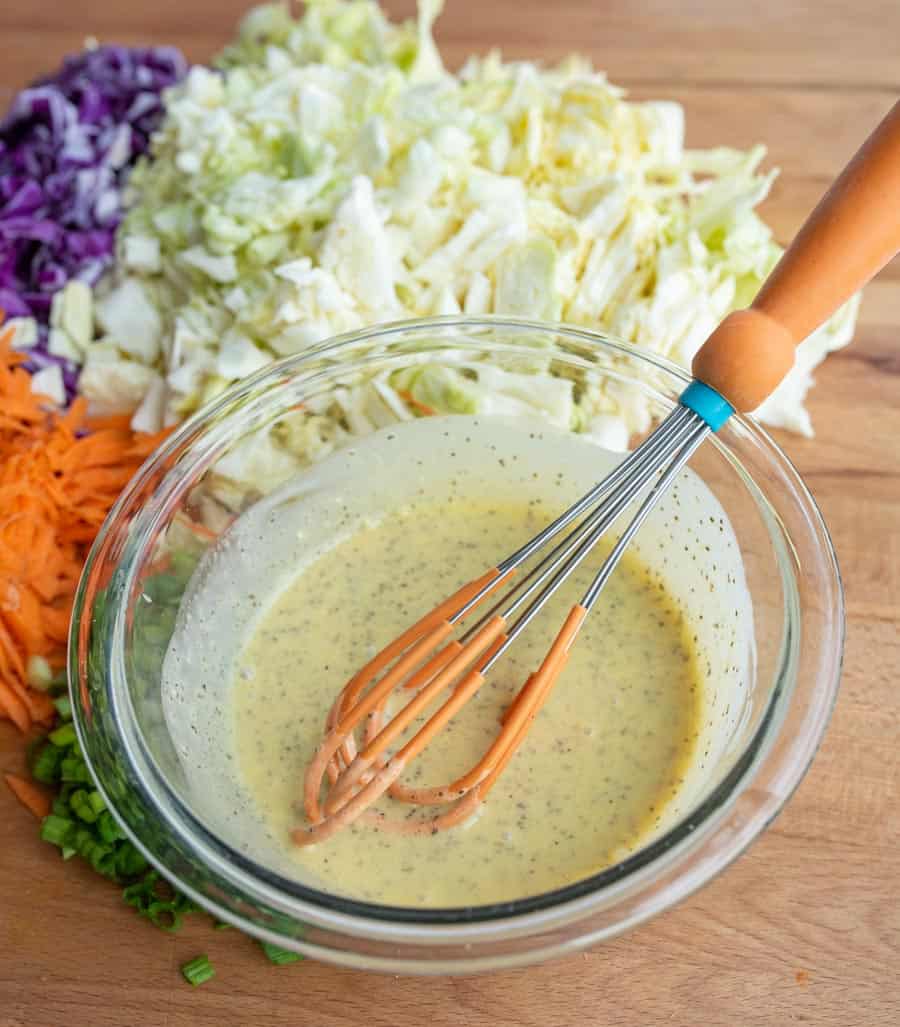 clear bowl with coleslaw dressing and a whisk in the bowl with all of the raw veggies around it including carrot - white and purple cabbage - and green onion