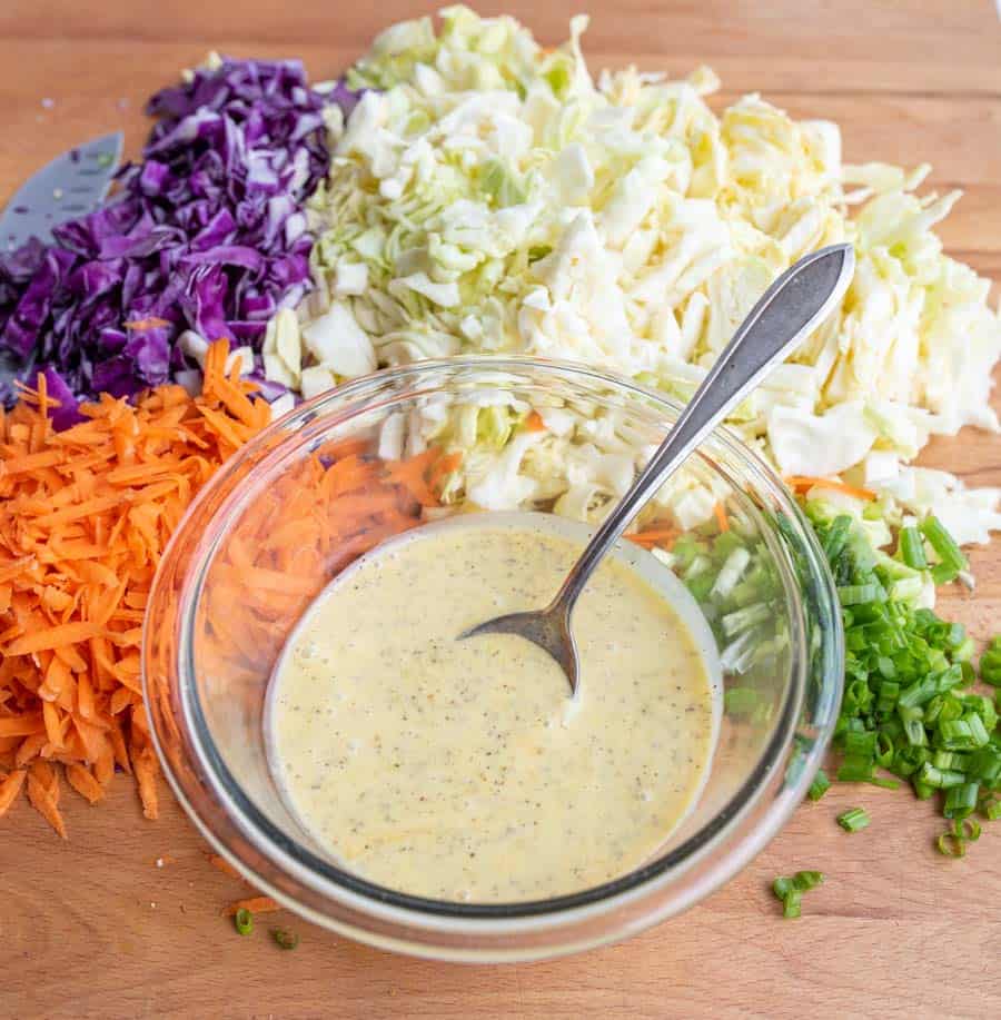 clear bowl with coleslaw dressing and a spoon with all of the raw veggies around it including carrot - white and purple cabbage - and green onion