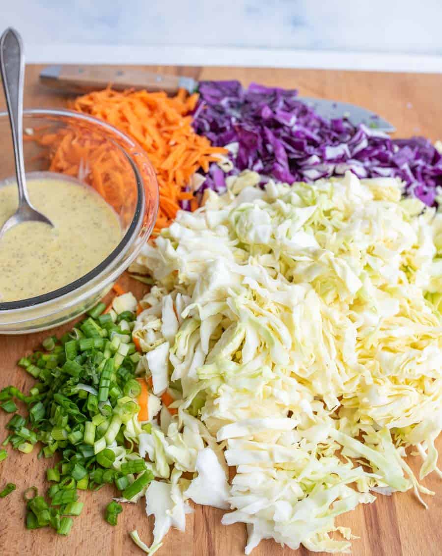 clear bowl with coleslaw dressing and a spoon with all of the raw veggies around it including carrot - white and purple cabbage - and green onion