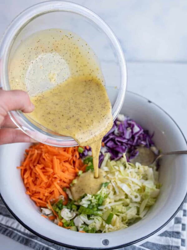 A bowl is being filled with the best coleslaw dressing recipe, poured from a smaller bowl. Inside the larger bowl are shredded carrots, purple cabbage, and green cabbage, with a spoon on the side. A striped cloth is underneath.