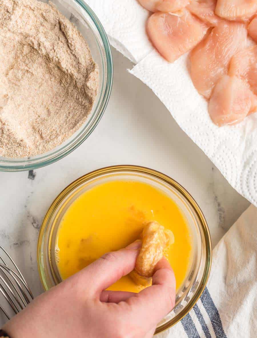 raw chicken being dipped in egg with the bowl of breading to the left.