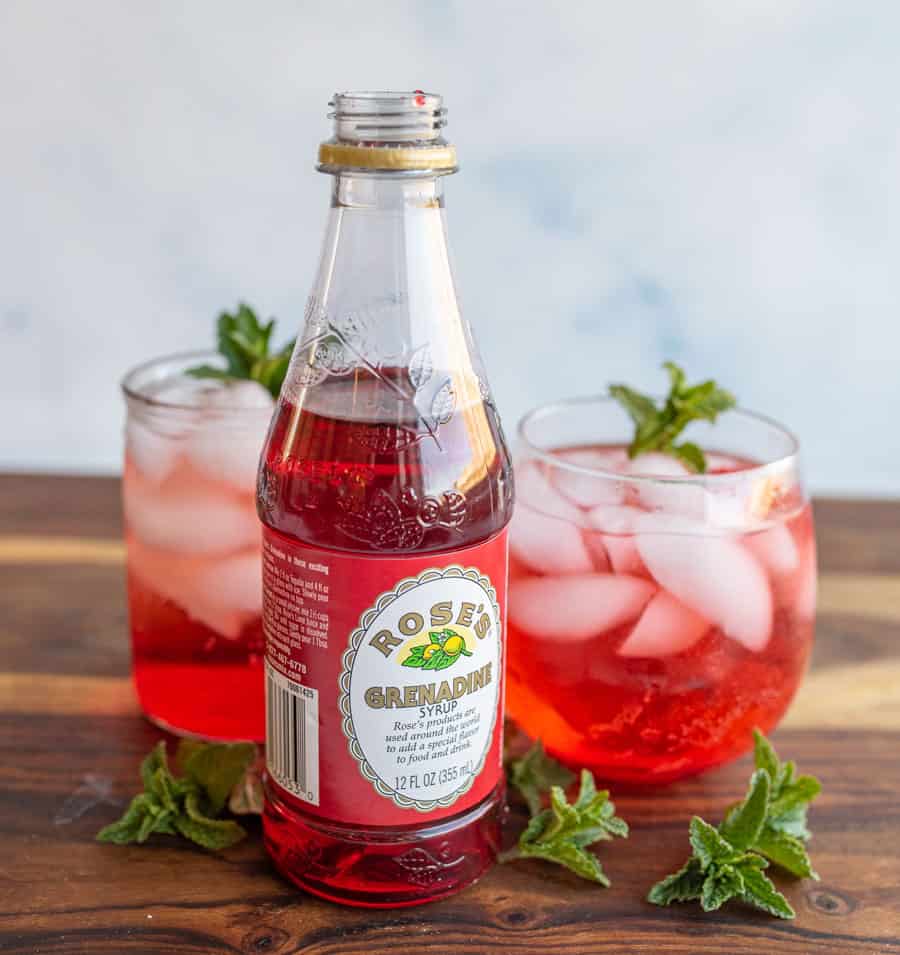 aesthetic photo of a bottle of 'Rose's Grenadine" with tumblers of ice and garnish and a red "Shirley-Temple" drink