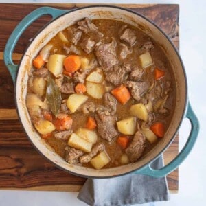 A pot of old fashioned beef stew recipe with chunks of beef, potatoes, carrots, and a bay leaf in brown broth sits on a wooden board with a gray cloth underneath.