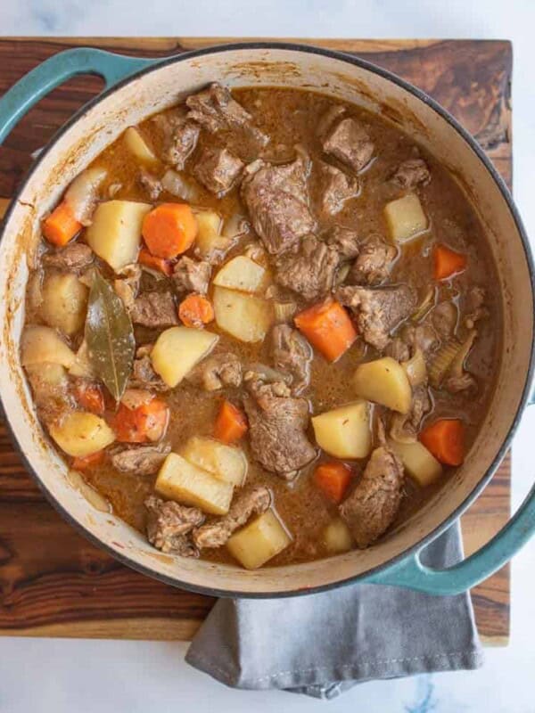 A pot of old fashioned beef stew recipe with chunks of beef, potatoes, carrots, and a bay leaf in brown broth sits on a wooden board with a gray cloth underneath.