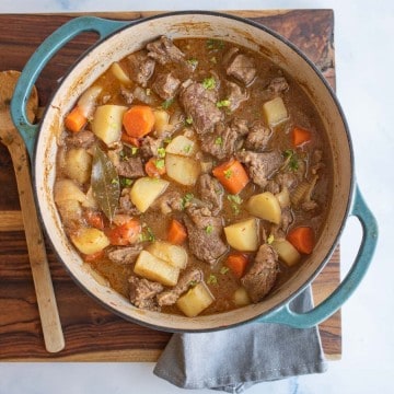 blue dutch oven with beef stew in it, looking from the top down, sitting on a cutting board garnished with chopped greens