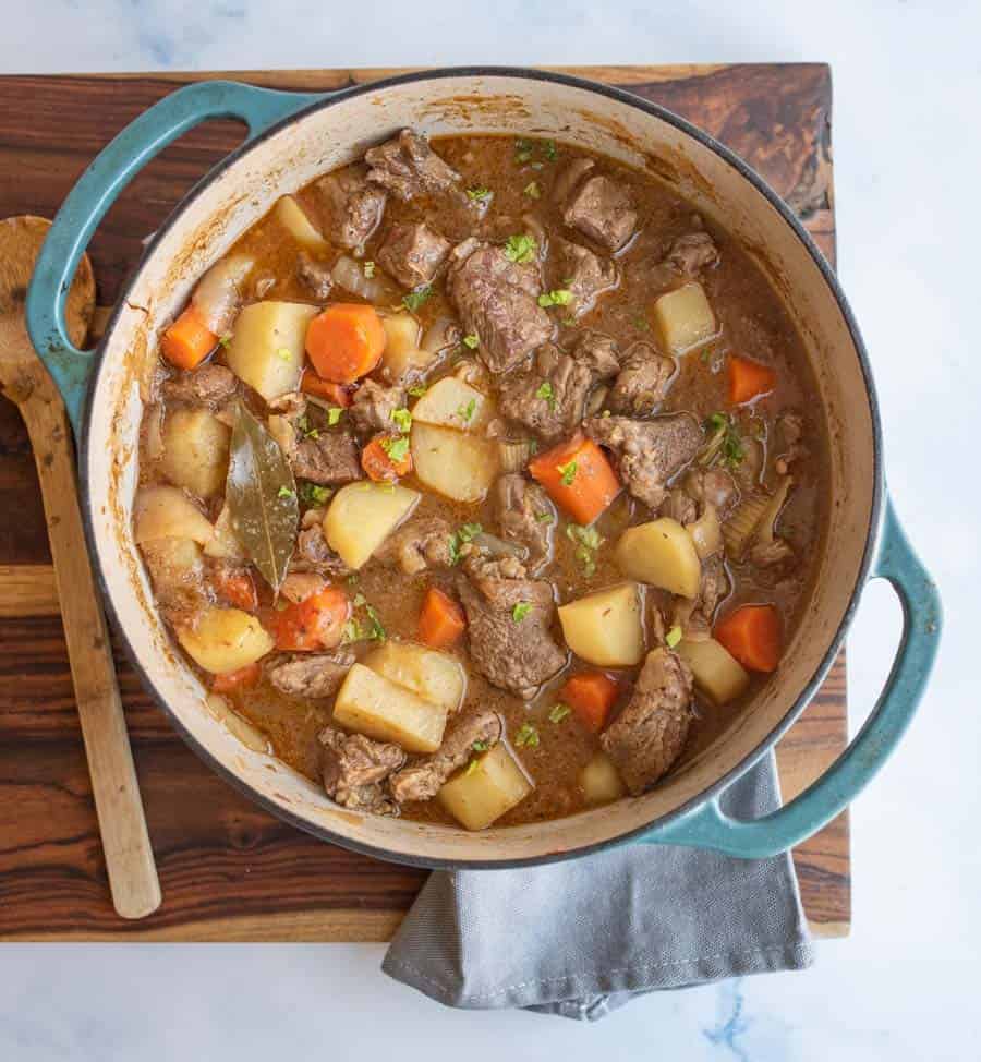 blue dutch oven with beef stew in it, looking from the top down, sitting on a cutting board garnished with chopped greens.