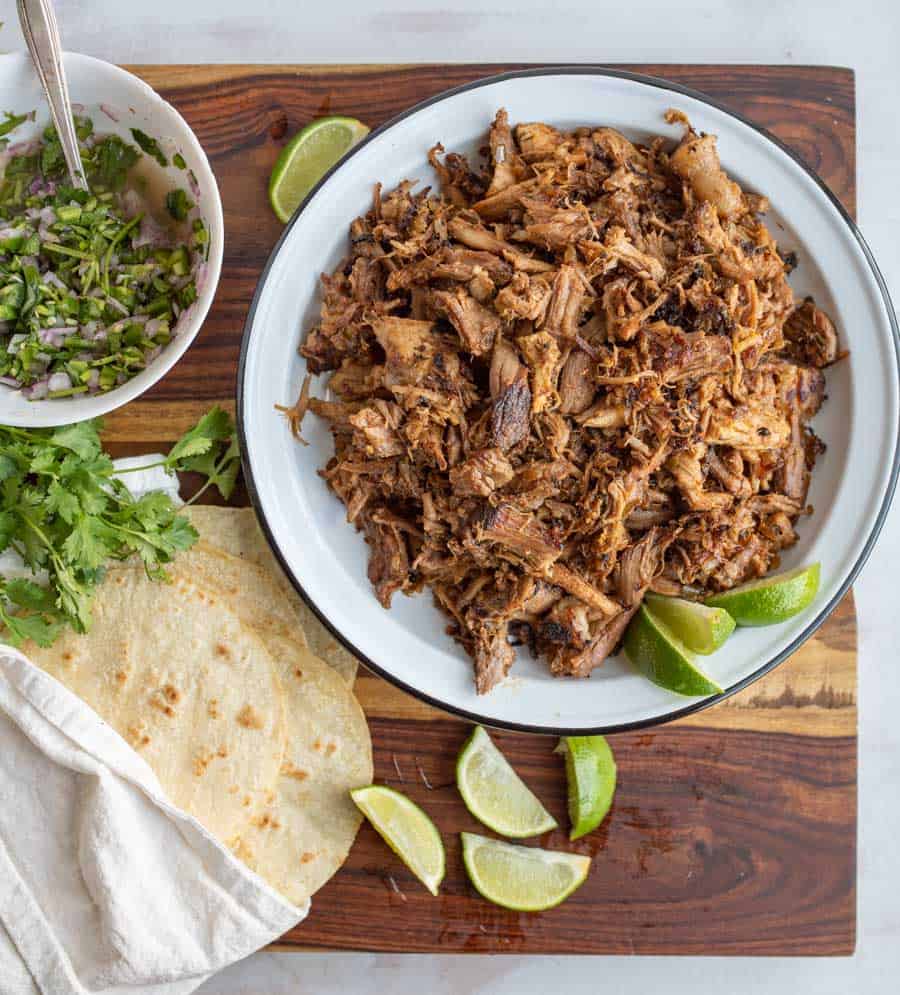 shreded cooked pork on a white plate with fresh lime with carnitas ingredients all around