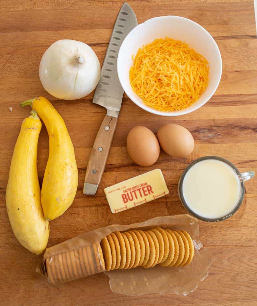 cutting board of ingredients for the squash casserole recipe consisting of summer squash cheese butter eggs onion crackers and a measuring cup of cream
