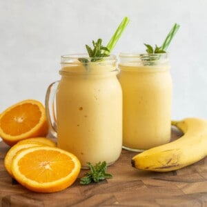 Two mason jars filled with creamy orange-yellow healthy tropical smoothies, garnished with mint and green straws, are on a wooden surface with sliced oranges, a whole banana, and sprigs of mint beside them.
