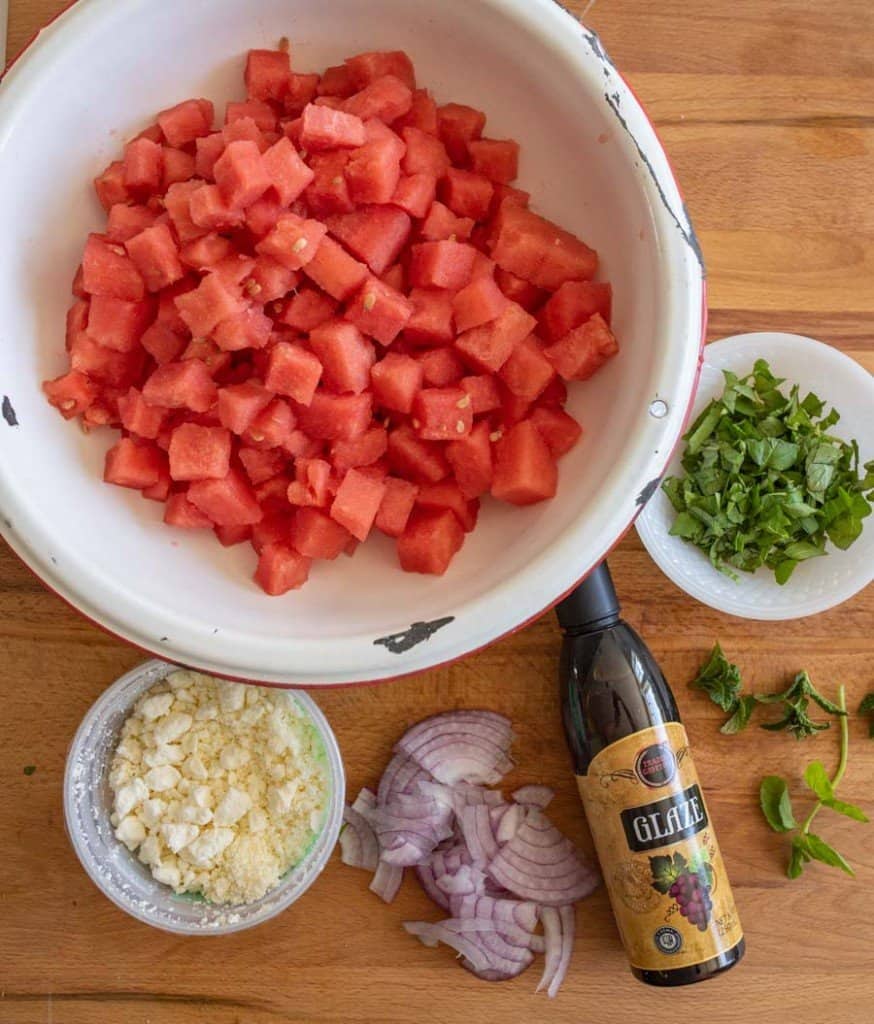 ingredients of watermelon and feta salad on a cutting board with around a big white bowl of chunked watermelon