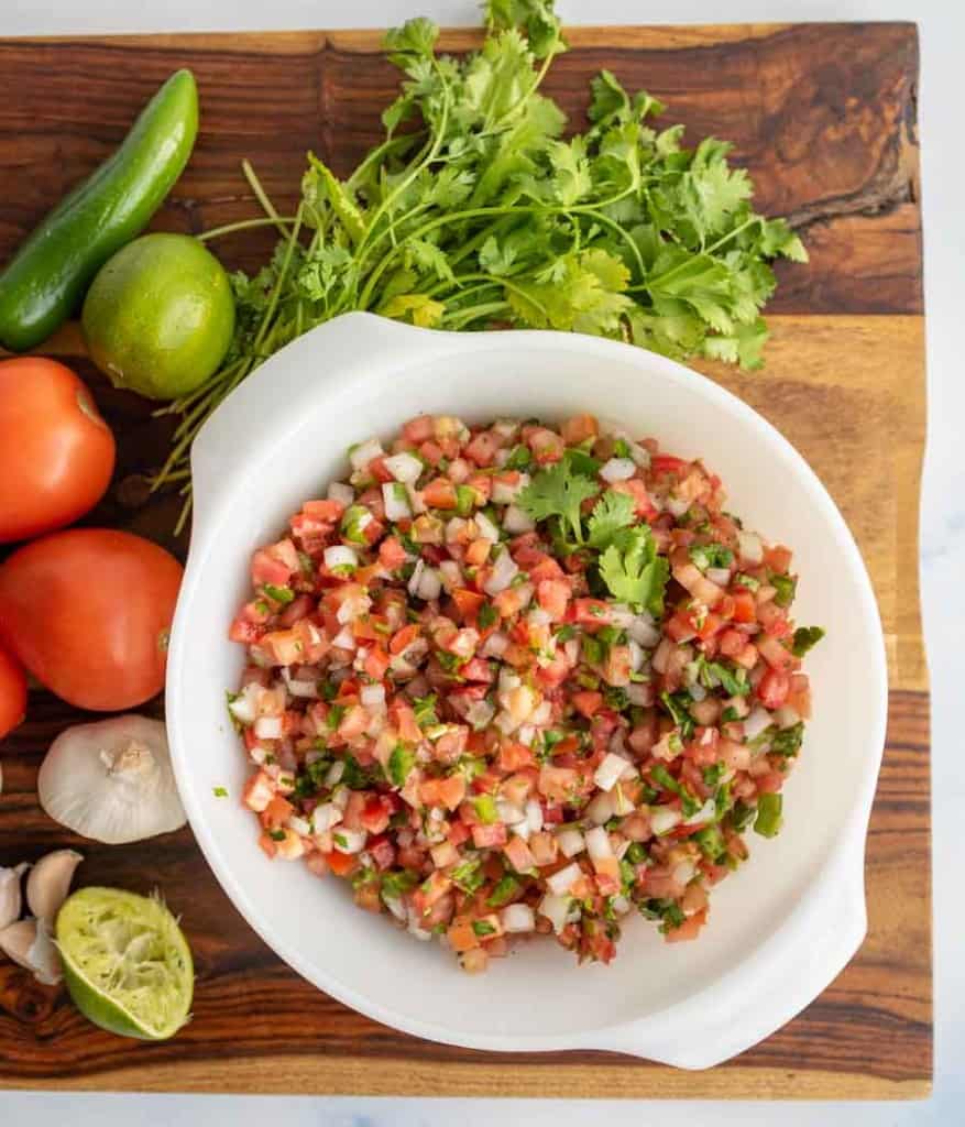 top view of a white bowl with chunky cut tomatoes, onion, herbs in a fresh pico de gallo surrounded by fresh ingredients