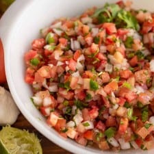 a white bowl with chunky cut tomatoes, onion, herbs in a fresh pico de gallo surrounded by limes