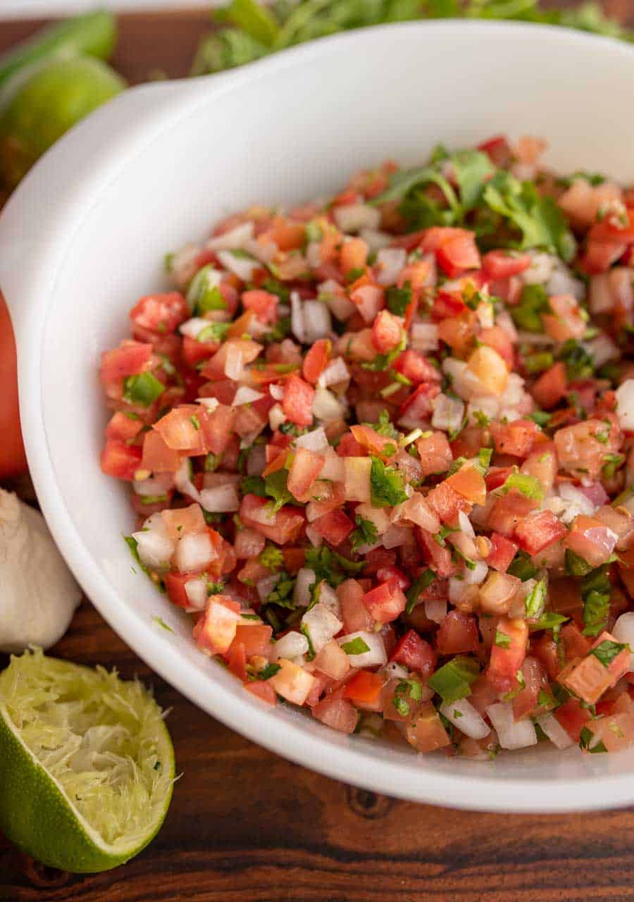 a white bowl with chunky cut tomatoes, onion, herbs in a fresh pico de gallo surrounded by limes