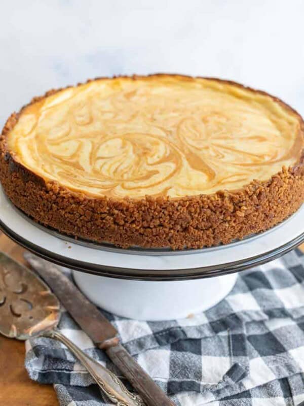 A marble-swirled cheesecake with a crumbly brown crust sits on a white cake stand. A pie server and knife rest nearby on a black and white checkered cloth on a wooden surface made from a pumpkin cheesecake recipe.