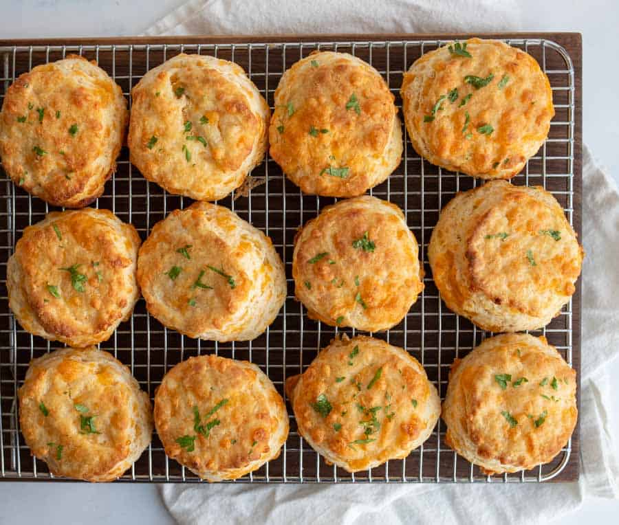 Twelve golden brown, round biscuits topped with chopped herbs are arranged neatly on a wire cooling rack over a white cloth.