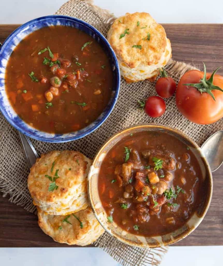 Two bowls of vegetable stew garnished with herbs, each served with biscuits on a wooden board. Fresh tomatoes and a spoon are nearby, and the board is set on a piece of burlap.