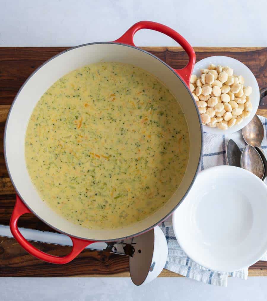 top view of red pot with cheddar broccoli soup with crackers, empty bowls, and a ladle off to the side