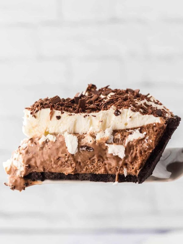 A slice of french silk pie with a chocolate cookie crust, creamy chocolate filling, whipped cream topping, and chocolate shavings on top, held up against a light, blurred background.