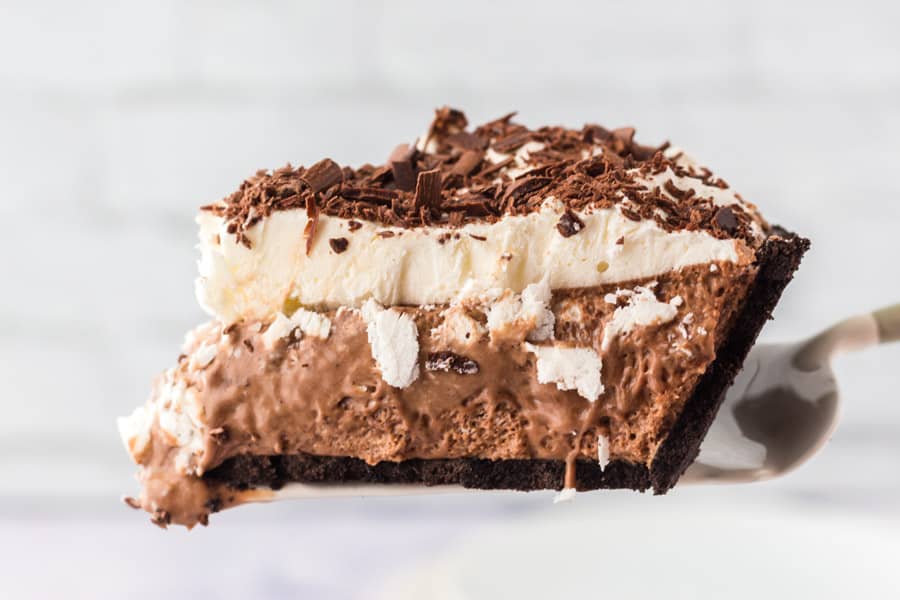 A close-up of a slice of chocolate cream pie with a chocolate crust, creamy filling, whipped topping, and chocolate shavings is held up on a spatula—perfect for dessert lovers who also enjoy treats like peach raspberry cobbler.