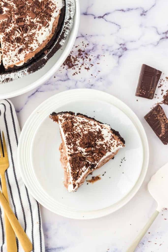 A slice of chocolate cream pie with whipped topping and chocolate shavings sits on a white plate next to a striped napkin, gold fork, and pieces of chocolate on a marble surface.