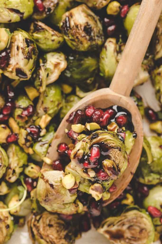 A wooden spoon scoops roasted Brussels sprouts in the oven, topped with pomegranate seeds and chopped pistachios, surrounded by more Brussels sprouts.