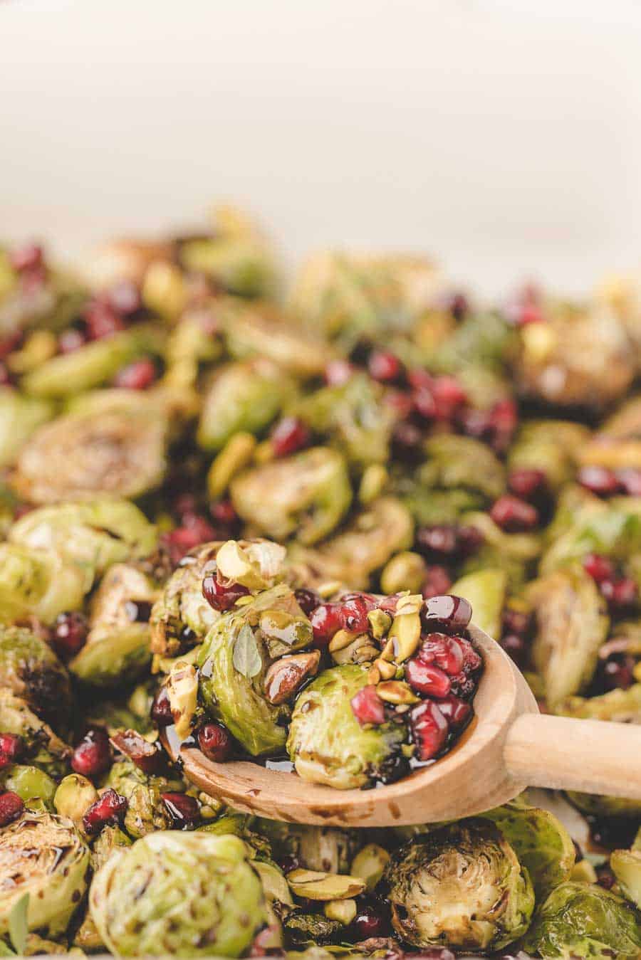 Oven-roasted Brussels sprouts mixed with pomegranate seeds and pistachios, served on a wooden spoon, with more of the roasted Brussels sprouts in the oven mixture in the background.