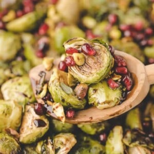 A wooden spoon holds roasted Brussels sprouts in the oven, topped with pomegranate seeds and pistachios, surrounded by more roasted Brussels sprouts in the background.