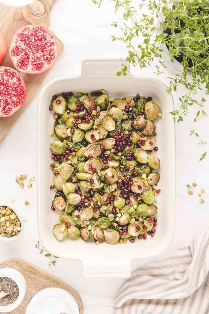 A white baking dish filled with roasted Brussels sprouts in the oven, pomegranate seeds, and pistachios sits surrounded by a halved pomegranate, fresh thyme, spices, and a striped towel on a white surface.
