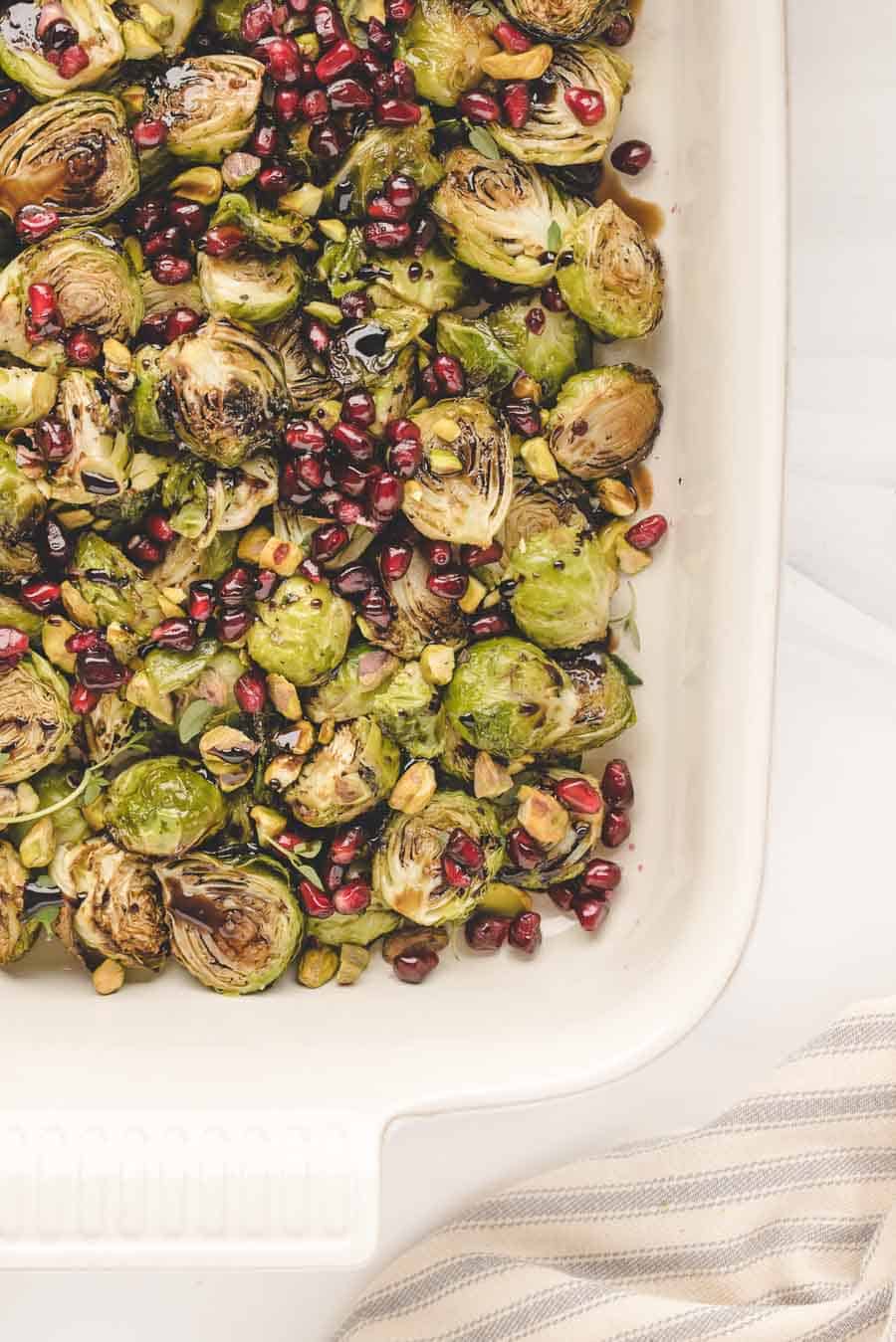 Roasted Brussels sprouts in the oven topped with pomegranate seeds, chopped pistachios, and a drizzle of balsamic glaze in a white baking dish on a light background.