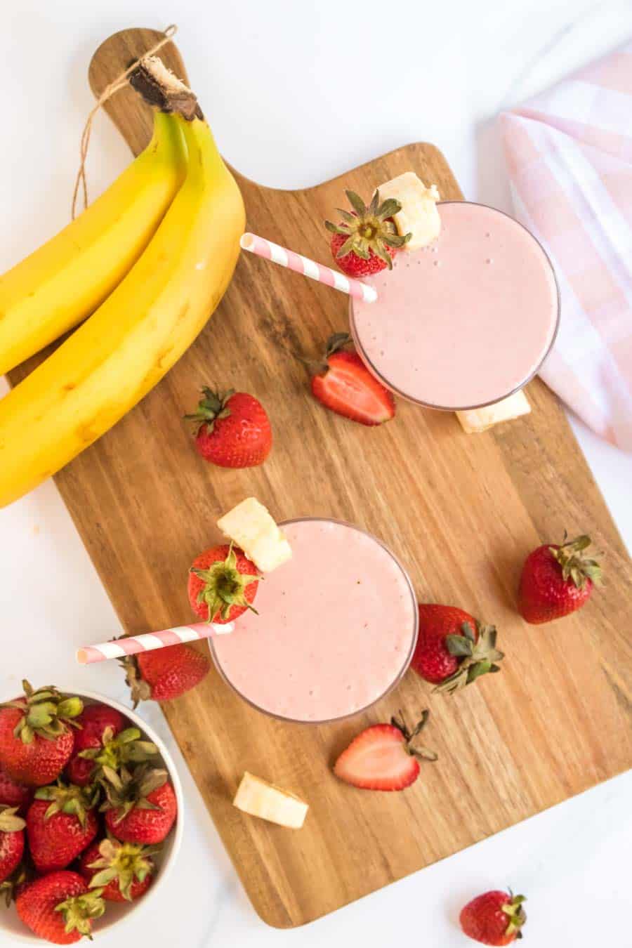 strawberry banana smoothies in cups with pink striped paper straws and fresh fruit all around