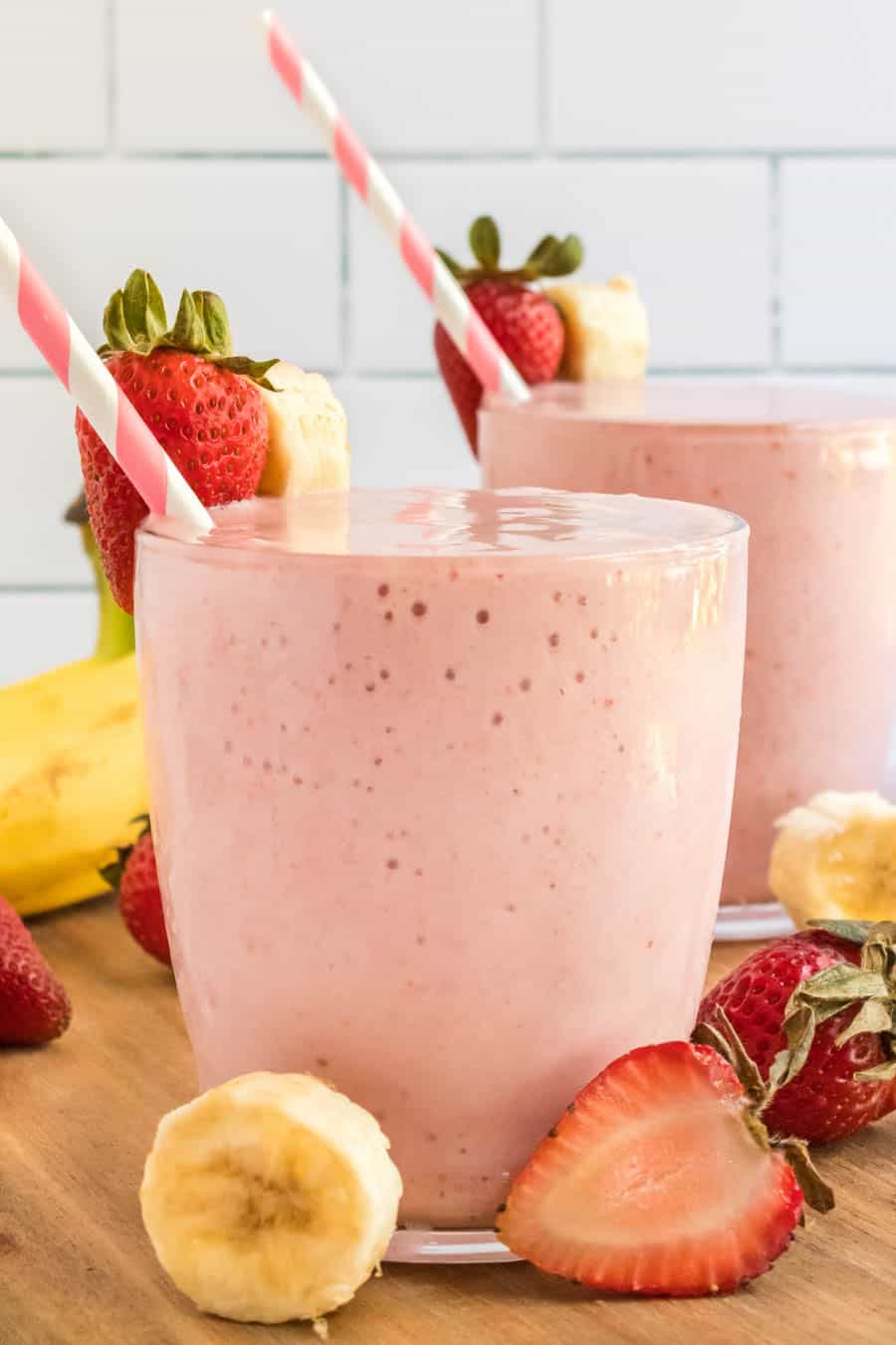 strawberry banana smoothies in cups with pink striped paper straws and fresh fruit all around