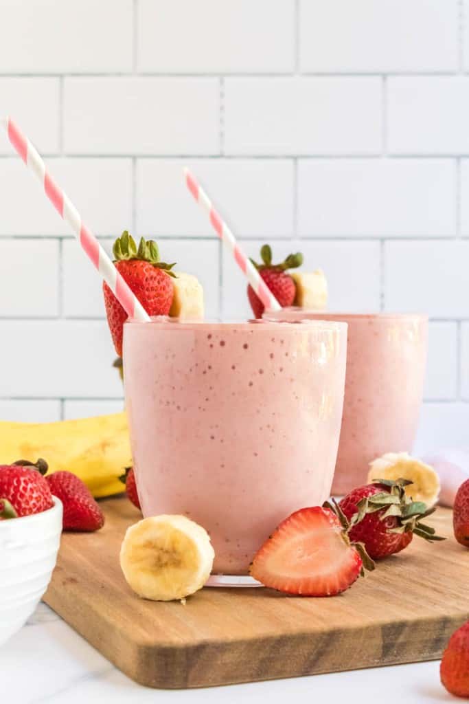 strawberry banana smoothies in cups with pink striped paper straws and fresh fruit all around