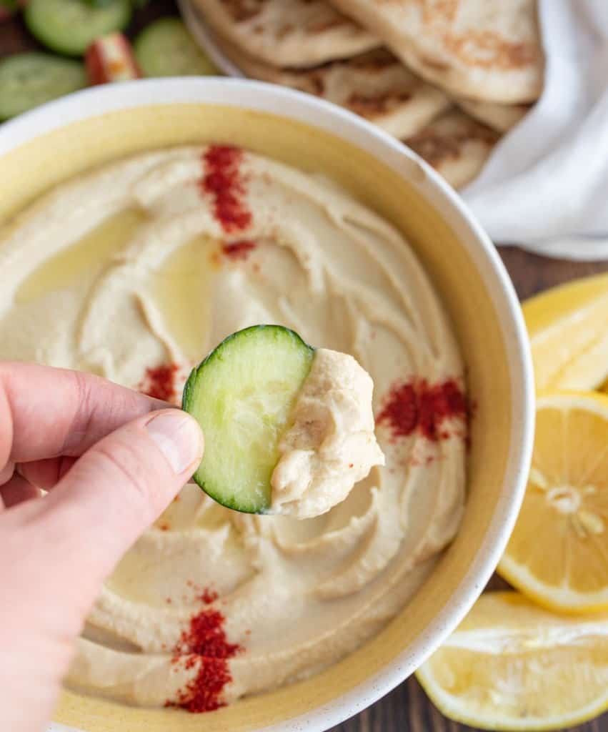 bowl of hummus with red spice on top POV scooping with slice of cucumber