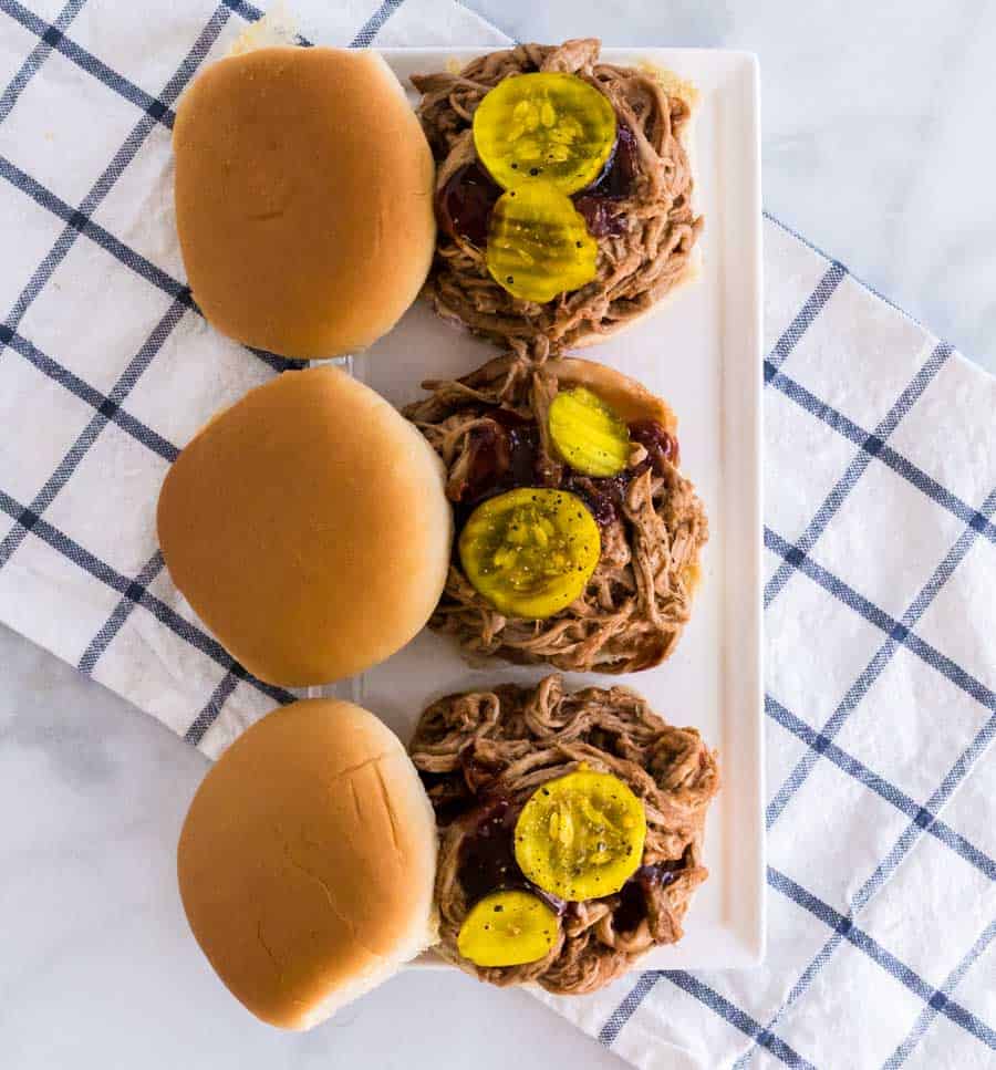 three pulled pork sandwiches in a row on a white plate over gingham cloth