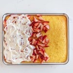 A rectangular cake, resembling an easy strawberry shortcake cake recipe, is partially topped with whipped cream and sliced strawberries on the left side. The right side remains plain. Served on a white dish, it sits elegantly atop a marble countertop.