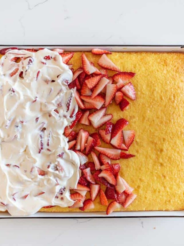 A rectangular cake, resembling an easy strawberry shortcake cake recipe, is partially topped with whipped cream and sliced strawberries on the left side. The right side remains plain. Served on a white dish, it sits elegantly atop a marble countertop.