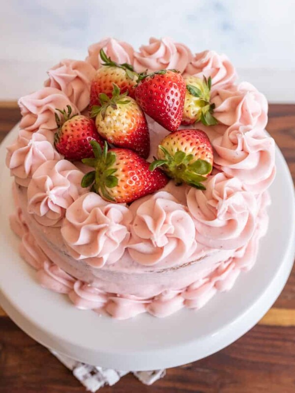 A round strawberry cake recipe with pink frosting and decorative swirls, topped with fresh whole strawberries, sits on a white cake stand on a wooden surface.