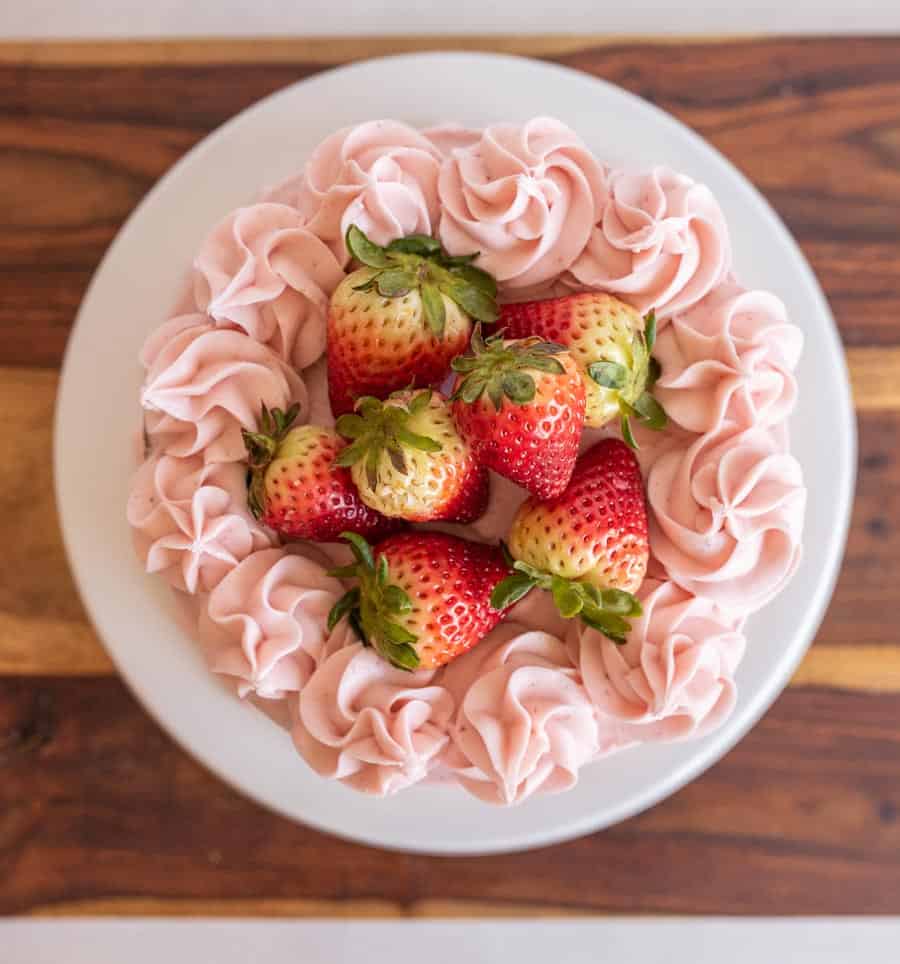 A round strawberry cake recipe topped with pink frosting swirls and whole strawberries, presented on a white plate and wooden surface.