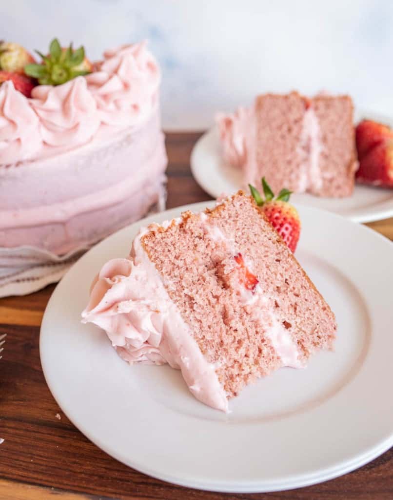 A slice of pink strawberry cake with strawberry frosting sits on a white plate, garnished with a fresh strawberry. The whole cake and another slice are visible in the background&mdash;perfect inspiration for your next strawberry cake recipe.
