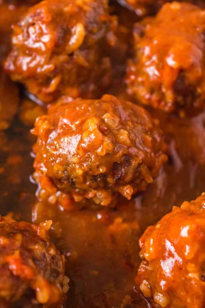 Close-up of several porcupine meatballs covered in a rich, reddish-brown tomato sauce, showing their moist and textured surface with visible pieces of onion and rice.
