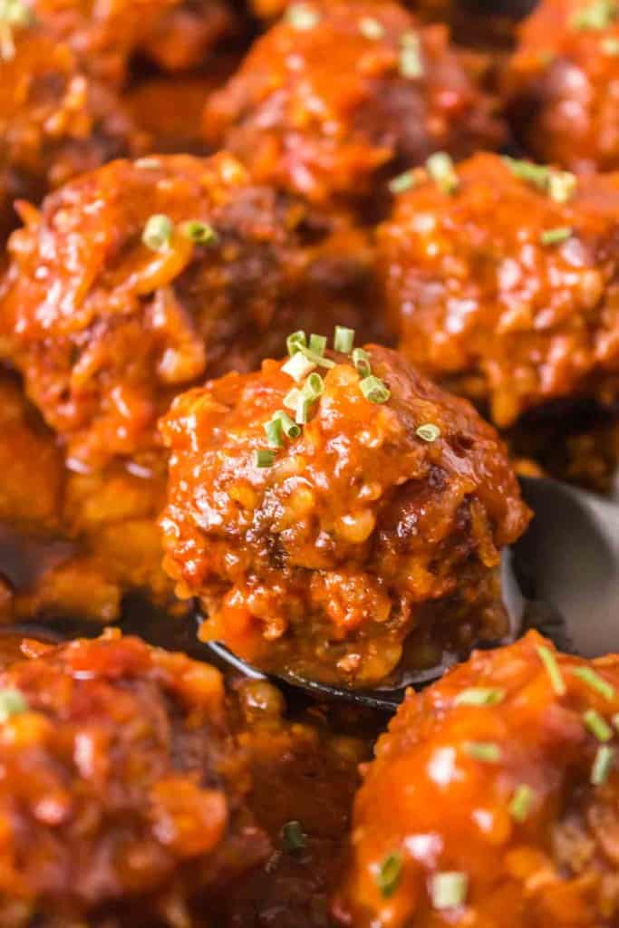 A close-up of saucy porcupine meatballs garnished with chopped green herbs, with one meatball being lifted from the pan on a spoon.