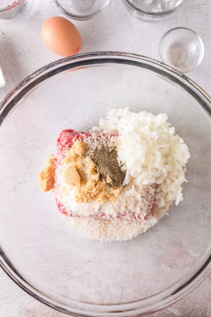 A glass bowl containing ground meat, chopped onions, uncooked rice, an egg, and seasonings&mdash;ingredients for porcupine meatballs&mdash;ready to be mixed. Surrounding the bowl are empty glass containers on a light surface.