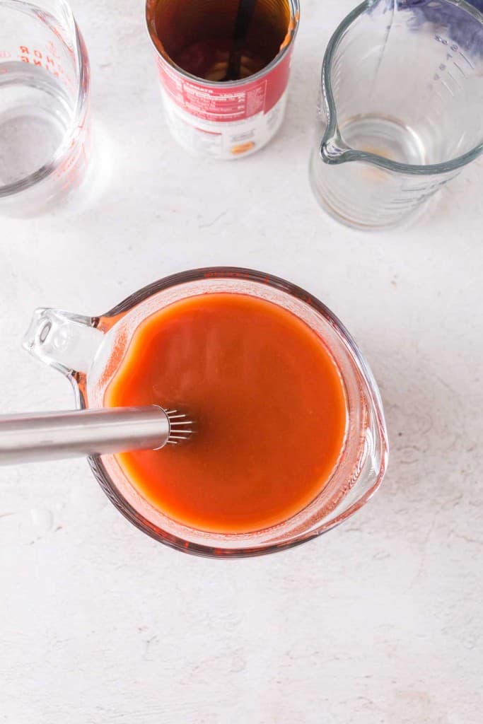 A metal whisk mixes a reddish-orange liquid in a glass measuring cup on a light surface&mdash;perfect for preparing sauce for porcupine meatballs&mdash;with an empty measuring cup, an open can, and a cup of water nearby.