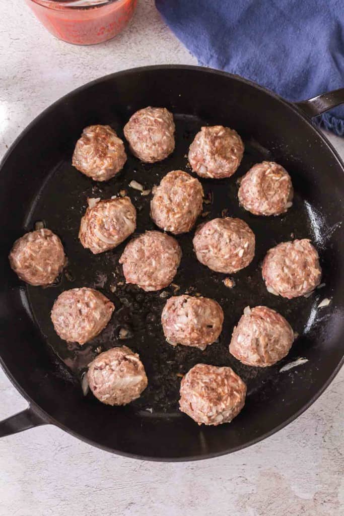 A black skillet with fourteen uncooked or lightly browned porcupine meatballs arranged inside, set on a light-colored surface next to a blue cloth and a partial view of a jar with red sauce.