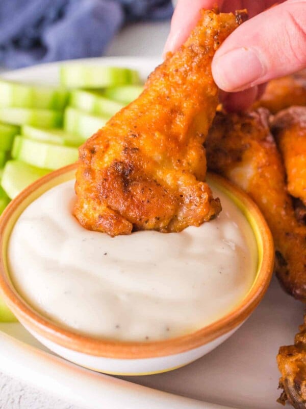 A close-up captures a hand dipping a golden-brown baked hot wings into a bowl of creamy white sauce. Sliced celery sticks sit invitingly in the background on a pristine white plate.