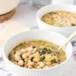 Two white bowls filled with white bean soup featuring greens and herbs. The soup is garnished with a sprinkle of cheese. A gold spoon rests in the foreground bowl. The setting includes a marble surface and a blurred background, highlighting the comforting dish.